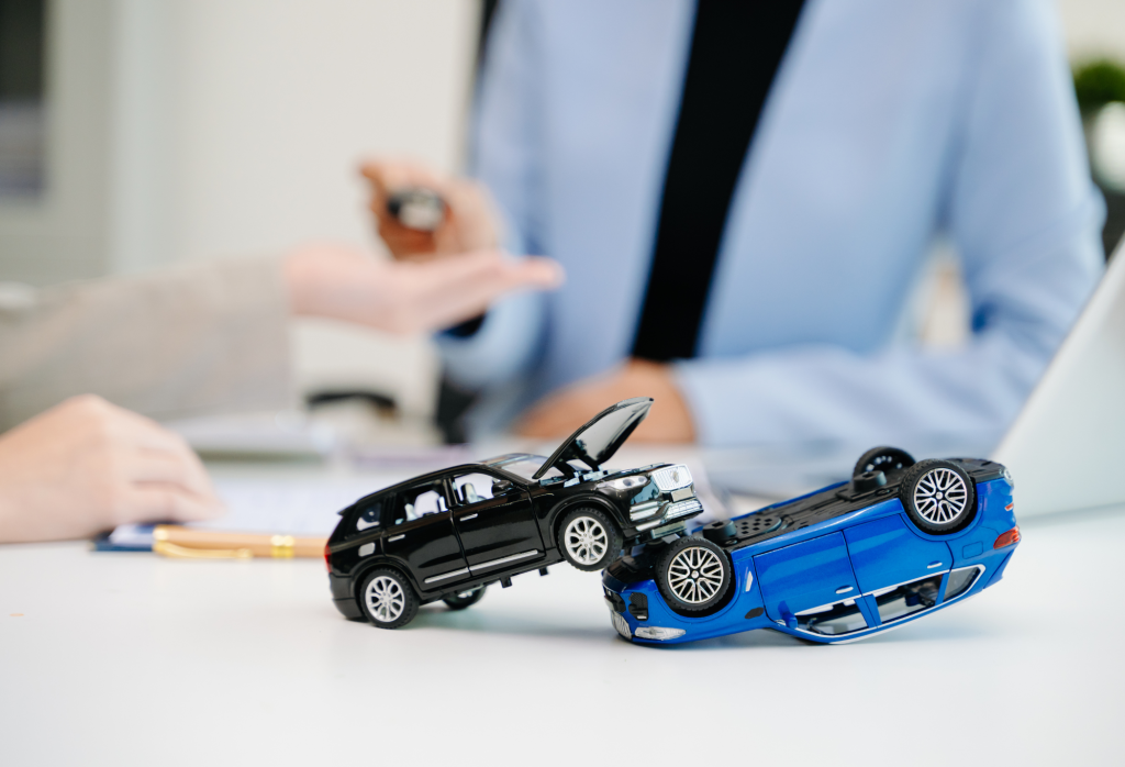 Close-up of two toy cars stacked on a blue truck model on a white table, with a blurred office scene in the background.